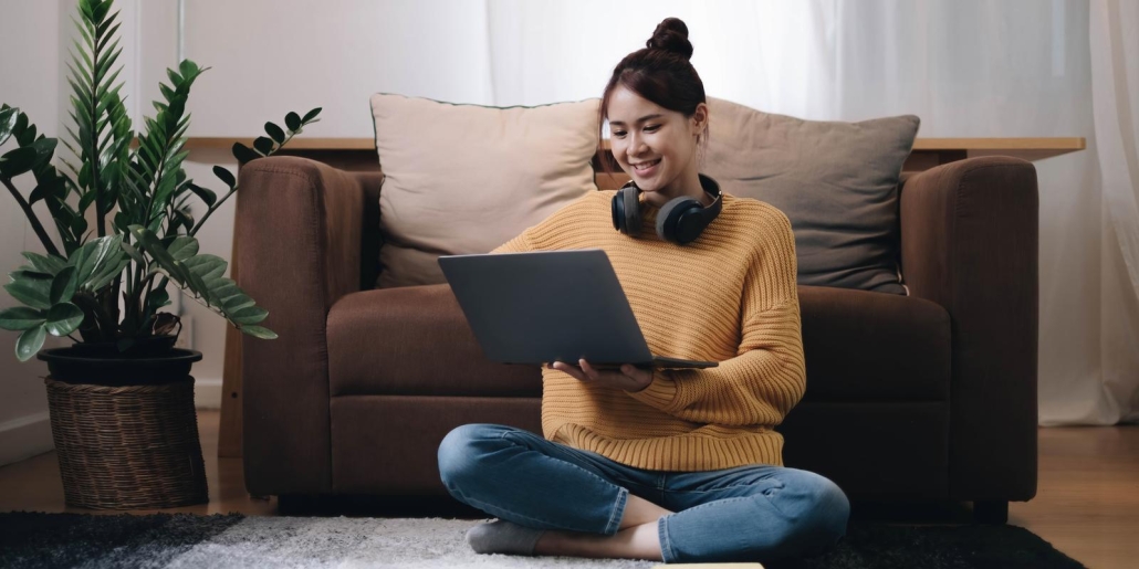 Smiling young woman working on laptop sitting on the floor in front of the sofa wearing headphones at home.
